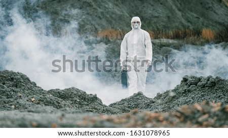 Lab technician in a mask and chemical protective suit, walks on dry ground with a tool box through toxic smoke.