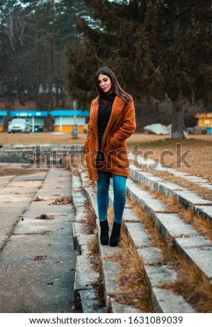 Portrait of beautiful brunette girl (woman) in orange fashion coat in season - photo on stairs with park and nature on background. Young female model posing and looking to camera - full body shot.