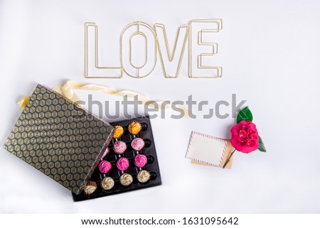 Top view festive composition of box with chocolate sweets with multicolored cream filling, wire word Love, blank retro postcard and rose flower on white background. Happy Valentine's day. Copy space