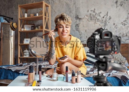 Young woman make-up blogger influencer sitting at stylish urban apartment recording video on digital camera applying bronzer on cheeks looking at mirror smiling happy