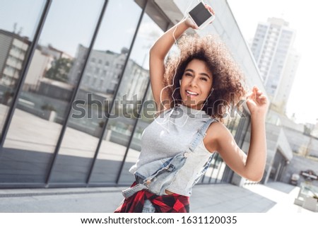 Young woman in the city street walking near window holding hand with smartphone up wearing earphones looking camera smiling happy close-up