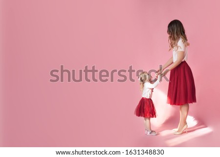 mother and her daughter in identical clothes hold white t-shirts and red tulle skirts from tulle and look at each other on a uniform pink background in a photo studio, mockup for women's day