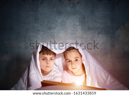Little girl and boy looking at each other under blanket. Children reading magic stories in bed before going to sleep. Young sister and brother in pajamas together on background of grey wall.