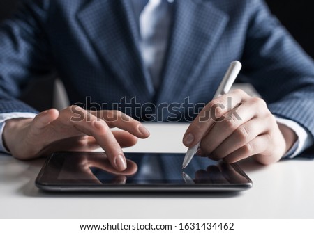 Man in business suit using digital tablet computer. Close-up of male hand holding pen and tablet device. Business man at workplace in office. Mobile smart device in business occupation.