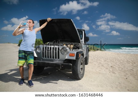 A caucasian tourist man drink bottle of water and looking on engine in car and tried deciding problem in the car on beach near Caribbean sea. 