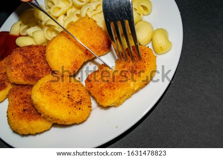 Fried chicken nuggets and boiled pasta with ketchup on a white plate on a dark background. Fork and knife cutting up the nuggets. Close up.