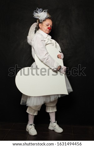 Serious woman in clown costume holding white empty heart banner on black background