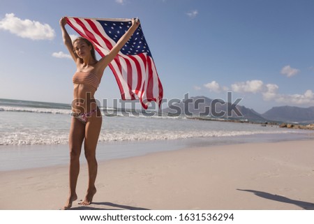Front view of young woman holding waving American flag at beach on a sunny day