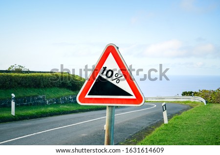 Red triangle road sign indicating a steep 10 percent downhill gradient in the road ahead. Empty road with crash barriers surrounded by green grass in the background. Traffic signs.