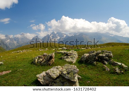 Large boulders protruding out of the lush green mountain top over looking a mountain range in the distance with sharp peaks disappearing into the clouds