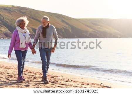 Loving Retired Couple Holding Hands As They Walk Along Shoreline On Winter Beach Vacation Royalty-Free Stock Photo #1631745025