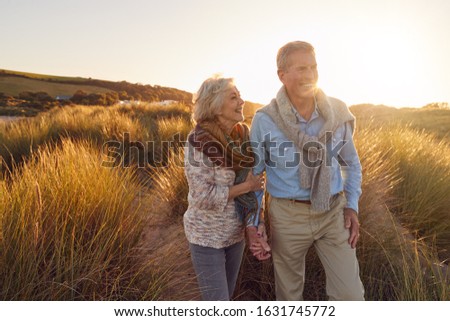 Loving Retired Couple Walking Arm In Arm Through Sand Dunes On Beach Vacation Against Flaring Sun Royalty-Free Stock Photo #1631745772