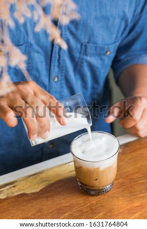 Male Barista pouring fresh milk froth on ice coffee for making an iced latte at the wooden counter bar.