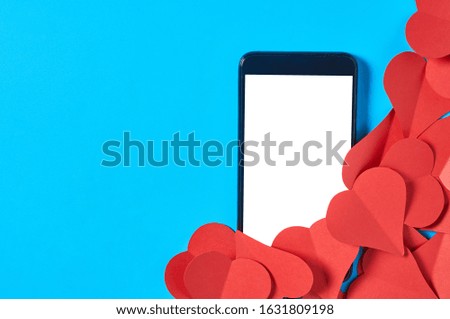 Black smartphone with isolated white screen for text, picture, photo and other graphics near heap of red paper hearts on blue table. Secrets or forbidden love concept. Space for text. Top view