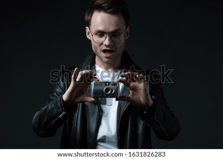 shocked stylish brutal man in biker jacket taking picture on film camera isolated on black
