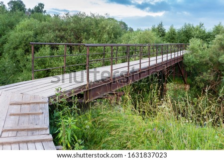 Old pedestrian bridge with a wooden flooring and an iron frame through a ravine in green thickets. Totma Russia. 