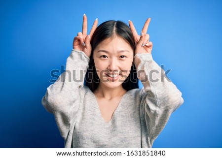 Young beautiful asian woman wearing casual sweater standing over blue isolated background Posing funny and crazy with fingers on head as bunny ears, smiling cheerful