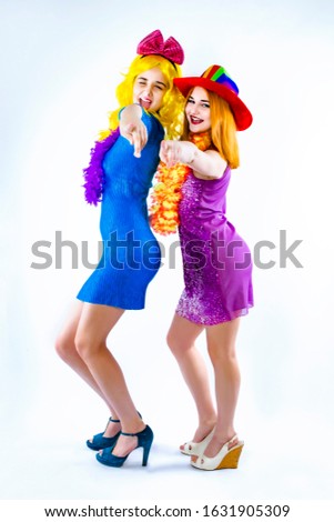 Two glamorous and beautiful girls in bright clothes and wigs have fun. Girlfriends posing while preparing for a Christmas party. Isolated on a light background.Point a finger at the camera