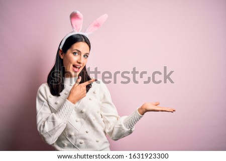 Young caucasian woman wearing cute easter rabbit ears over pink isolated background amazed and smiling to the camera while presenting with hand and pointing with finger.