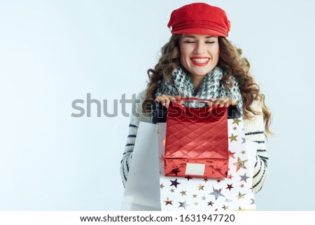 happy trendy middle age woman with long brunette hair in sweater, scarf and red hat with shopping bags isolated on winter light blue background.