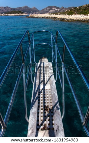 A picture taken from the bow of a sailing boat and white wooden gangway with shiny metal handles can be seen over the crisp turquoise waters of Mediterranean coast of Kas, Turkey.