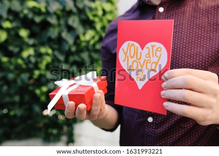 Young man holding valentine card and pretty red present box in the hands. Celebrating Happy Valentine`s day. February 14. Surprise . Love. Concept of Valentines Day. Object