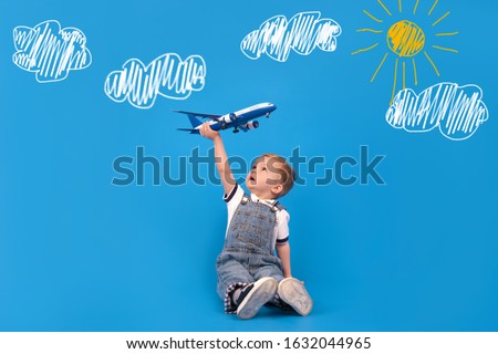 Happy child sits holding plane in his hand and imagining how he is flying on plane on blue background and drawings of white clouds and yellow sun. Imagination, creativity and concept ideas.