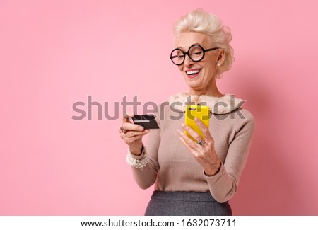 Grandmother makes a payment, using a credit card and smartphone. Photo of kind elderly woman on pink background