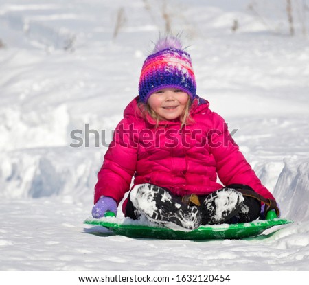 Young child, having fun, sledding on saucer like object, down snowy hillside in winter.