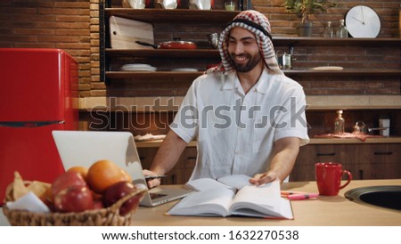 Smiling handsome young arabic man use laptop look up in book write at table kitchen successful hijab browsing business emirates ethnic home modern work confident internet muslim portrait slow motion