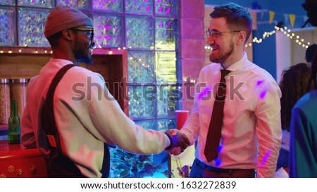 On weekend party, happy young man turning on old record player enjoying music shaking hands with best friend hanging out together at night.