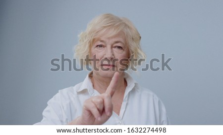 Cheerful blonde old woman using a virtual technology screen in air pointing with fingers and smiling of joy on light background.