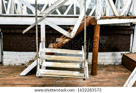An old wooden swing, painted white and suspended from ropes. The white paint was peeling.  Old wooden terrace with stairs in the background