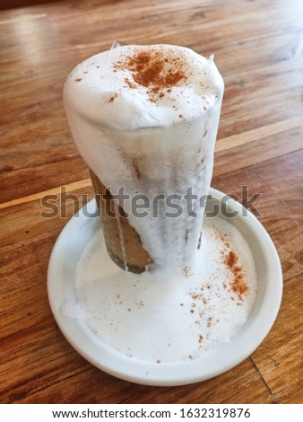 Iced coffee with frothy milk overflowing glass topping with powder of cinnamon, cuppuchino trendy style serving in cafe, restaurant, vertical picture tall glass over white plate on wooden table 