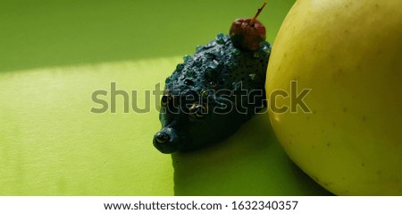 A plasticine hedgehog in small beads looks out from behind a wet yellow Apple on a green sheet of paper.