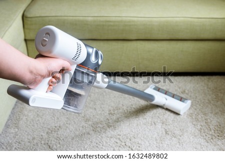 Wireless vacuum cleaner used on carpet in room. Housework with new handy upright stick hoover. Person holds vacuum cleaner, modern white broom by sofa and floor. Home cleaning and care concept. Royalty-Free Stock Photo #1632489802