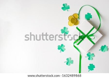 Composition for St. Patrick's Day. Gift white box with a green satin bow, clover and toy gold coins on a white background. The view from the top, lay flat, copy space