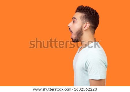 Side view portrait of surprised brunette man with beard in white t-shirt looking left with big eyes and open mouth, shocked by news, empty copy space for text. studio shot isolated, orange background Royalty-Free Stock Photo #1632562228