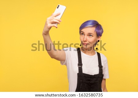 Portrait of stylish trendy hipster girl with violet hair, modern haircut and in denim overalls holding smartphone and taking selfie photo, making video call. isolated on yellow background, studio shot