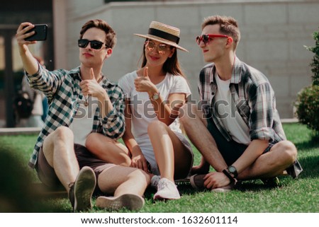 Three Light-Skinned Guys Pose For Selfies Sitting On The Lawn On A Summer Day. Two Attractive Boys And A Girl In A Hat And Sunglasses Take Pictures And Relax On Vacation.