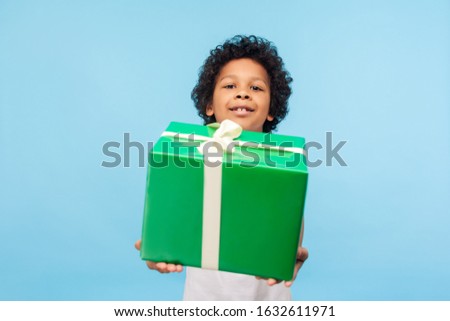 Take this present! Happy generous good-natured little boy with curly hair giving gift box and looking at camera with toothy smile, holiday charity. indoor studio shot isolated on blue background