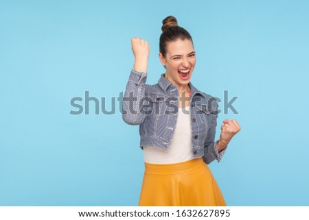 Portrait of excited overjoyed fashionably dressed woman with hair bun standing with raised fists and shouting yeah, I'm winner, rejoicing victory, success. studio shot isolated on blue background Royalty-Free Stock Photo #1632627895
