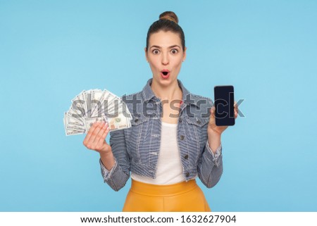 Unbelievable mobile payment service. Portrait of surprised woman with hair bun holding cellphone and dollar bills, looking at camera with amazement shock. studio shot isolated on blue background