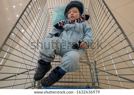 funny little boy in blue ski suit and striped scarf lies and sleeps in grocery cart from store