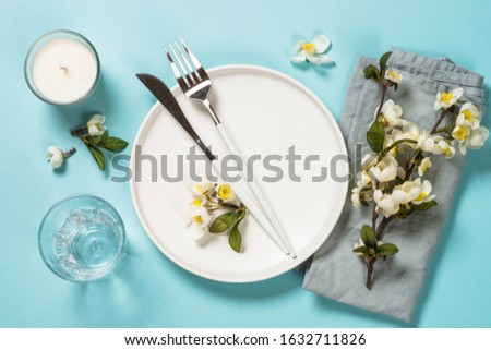 Spring table setting with blossom flower, white plate and cutlery on blue background. Flat lay image.