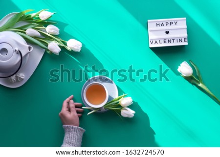 Spring geometric flat lay. Female hands hold lightbox with text "Happy Valentine". Tea cup, pot, sweets and white tulips on green. Valentine's day, sunlight, long shadow design, top view