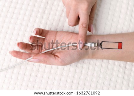 Low battery symbol drawn on human hand. Tired woman lying on the bed, trying to charge from USB, holding cable with your finger. Overwork, exhausted, chronic fatigue, living energy concept.  Royalty-Free Stock Photo #1632761092