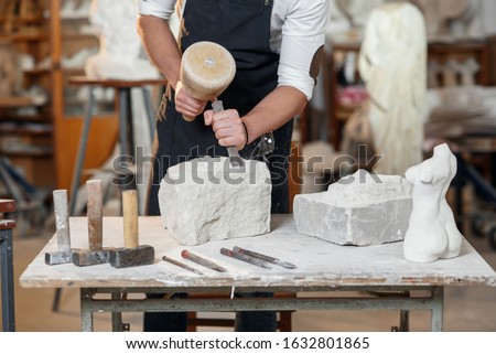 Bearded craftsman works in white stone carving with a chisel. Creative workshop with works of art. Royalty-Free Stock Photo #1632801865