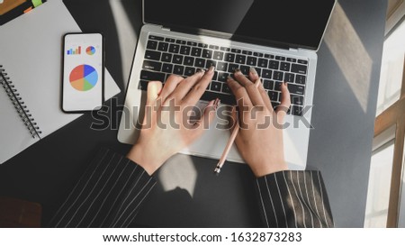 Top view of businesswoman working on laptop while looking business chart on smartphone with other office supplies in comfortable workplace