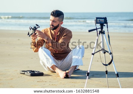 arabian man sitting on sand and talking tripod video making review of quadrocopter in beach. Travel videographer lifestyle content maker concept